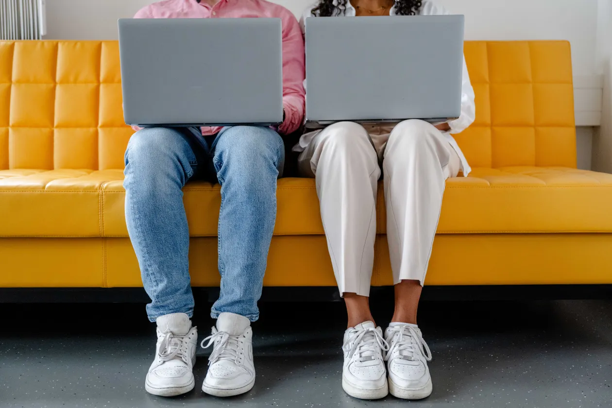 Foto: Zwei Personen sitzen nebeneinander und haben jeweils einen aufgeklappten Laptop auf dem Schoß. Man sieht nur ihre Beine und die Laptops.