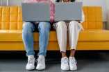 Photo: Two people sitting next to each other, each with an open laptop on their lap. You can only see their legs and the laptops.