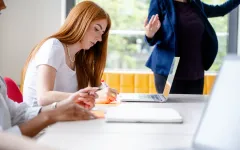 Das Bild zeigt eine junge Frau in einer Workshopsituation, vor sich ein Laptop macht sie Notizen, hinten schemenhaft die Vortragende.