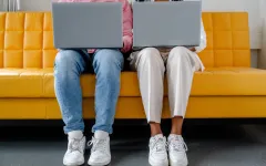 Foto: Zwei Personen sitzen nebeneinander und haben jeweils einen aufgeklappten Laptop auf dem Schoß. Man sieht nur ihre Beine und die Laptops.