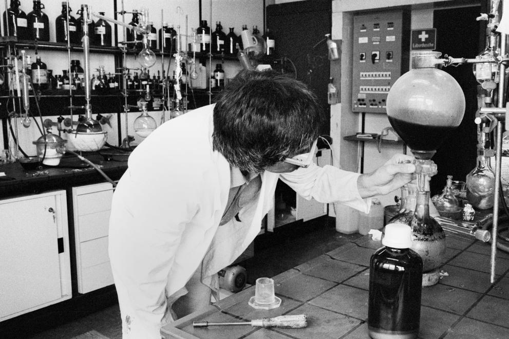 Researcher in a chemistry laboratory in 1983, the picture shows many glass flasks and a shelf with many bottles.